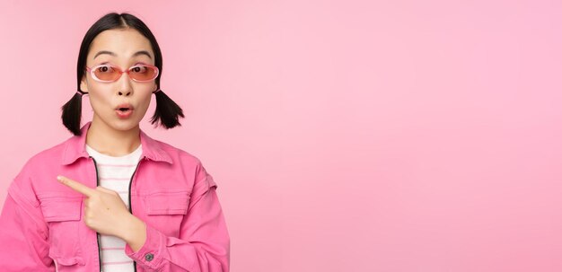 Retrato de una chica asiática sonriente con elegantes gafas de sol apuntando con el dedo a la izquierda mostrando una pancarta publicitaria de pie sobre un fondo rosa