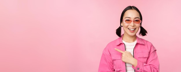 Retrato de una chica asiática sonriente con elegantes gafas de sol apuntando con el dedo a la izquierda mostrando una pancarta publicitaria de pie sobre un fondo rosa