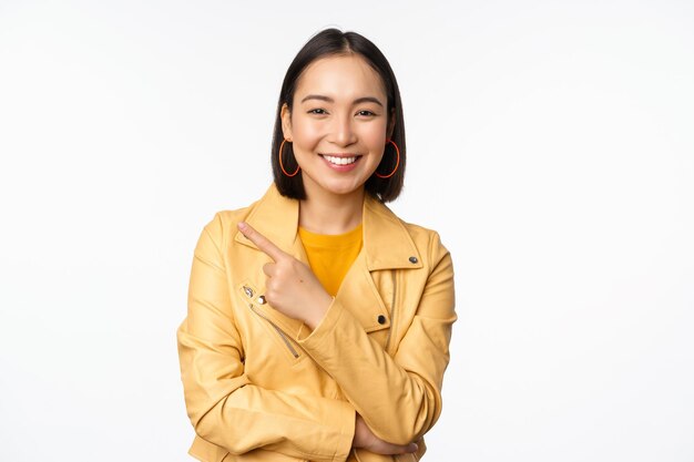 Retrato de una chica asiática sonriente que señala con el dedo a la izquierda mostrando el logotipo o el anuncio del espacio de copia en el lado vacío de pie sobre fondo blanco