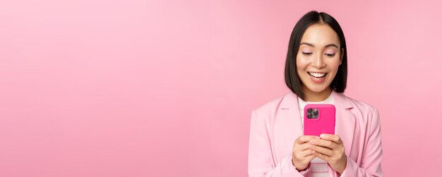 Retrato de una chica asiática en traje con teléfono inteligente sonriendo y luciendo feliz de pie sobre fondo de estudio rosa