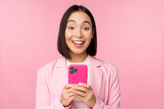 Retrato de una chica asiática en traje con teléfono inteligente sonriendo y luciendo feliz de pie sobre el fondo rosa del estudio Copiar espacio