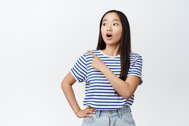 Retrato de una chica asiática se ve sorprendida, dice wow señalando con el dedo a la izquierda en la promoción de venta que muestra un banner de descuento sobre fondo blanco