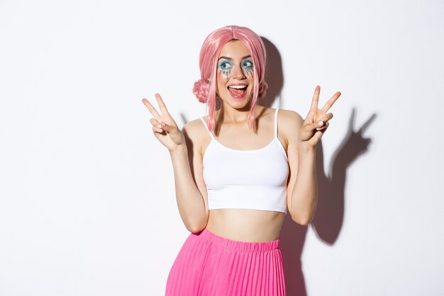 Retrato de chica atractiva elegante con peluca rosa y disfraz de halloween, mostrando signos de paz y sonriendo feliz, de pie.