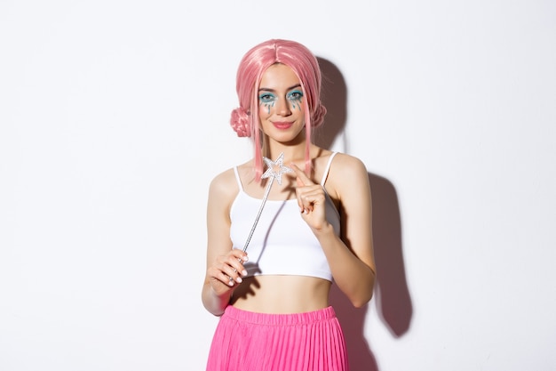 Retrato de una chica atractiva con peluca rosa y maquillaje brillante, vestida como un hada para la fiesta de halloween, sosteniendo la varita mágica y sonriendo