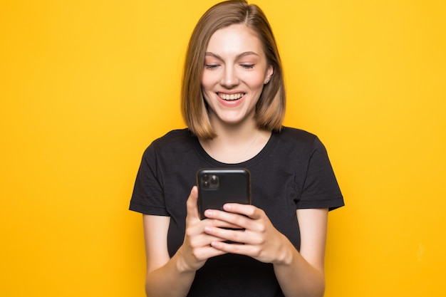 Retrato de chica atractiva sosteniendo un teléfono inteligente en las manos, revisando el correo electrónico, usando internet 5G, escribiendo sms, de pie sobre una pared amarilla
