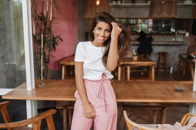 Retrato de una chica bronceada con camiseta blanca y pantalones rosas posando en un café con muebles de madera