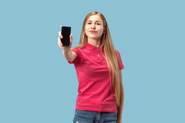Foto gratuita retrato de una chica casual confidente que muestra el teléfono móvil de la pantalla en blanco aislado sobre la pared azul