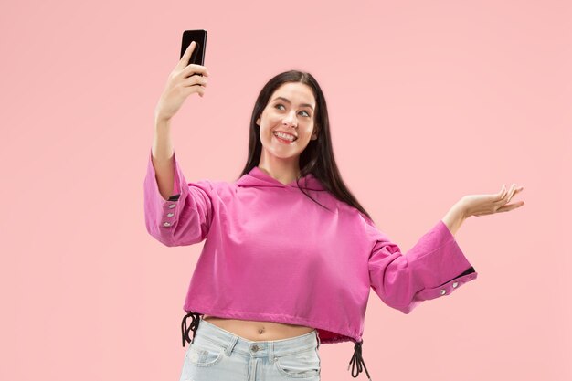 Retrato de una chica casual sonriente feliz confiada que hace la foto del selfie por teléfono móvil aislado sobre la pared rosada.