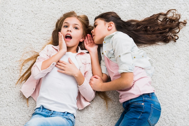 Retrato de una chica chismosa contando un secreto en el oído a su amiga tendida en la alfombra