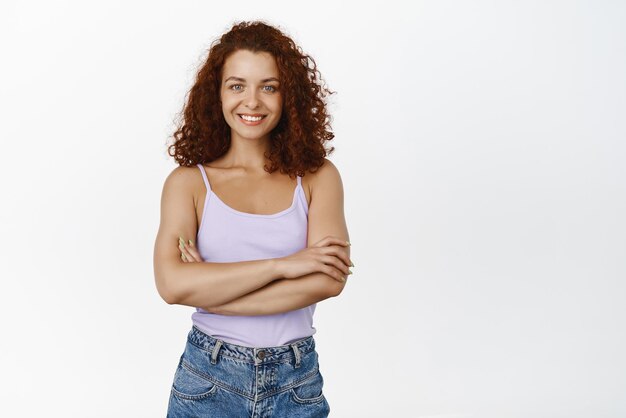 Retrato de una chica confiada en forma y con estilo con el pelo rojo rizado cruza los brazos en el pecho y se ve confiada motivando la expresión segura de sí misma fondo blanco