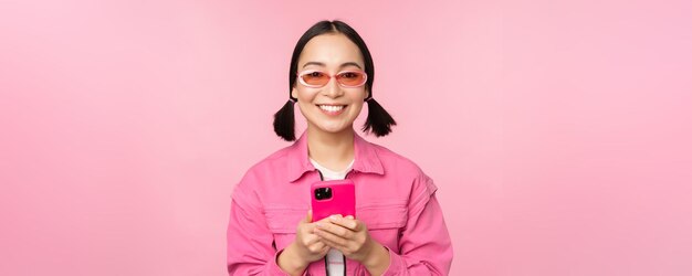 Retrato de una chica coreana con elegantes gafas de sol sosteniendo un teléfono móvil usando una aplicación de teléfono inteligente de pie