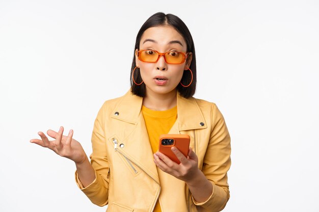 Retrato de una chica coreana con gafas de sol sosteniendo un teléfono inteligente mirando confundido y encogiéndose de hombros de pie sobre fondo blanco