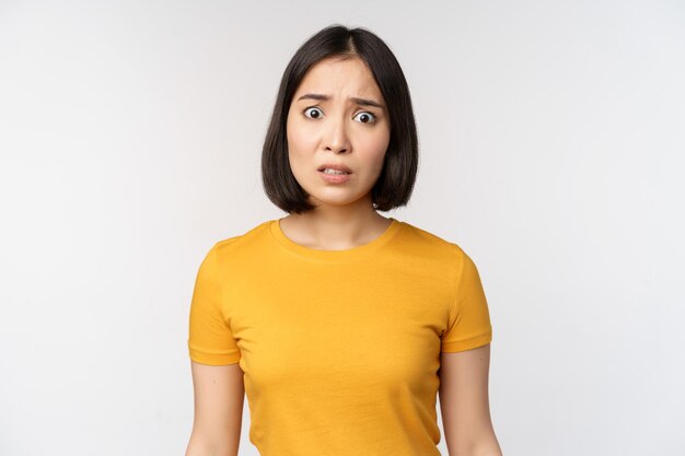 Retrato de una chica coreana preocupada mirando preocupada a la cámara de pie con una camiseta amarilla sobre fondo blanco