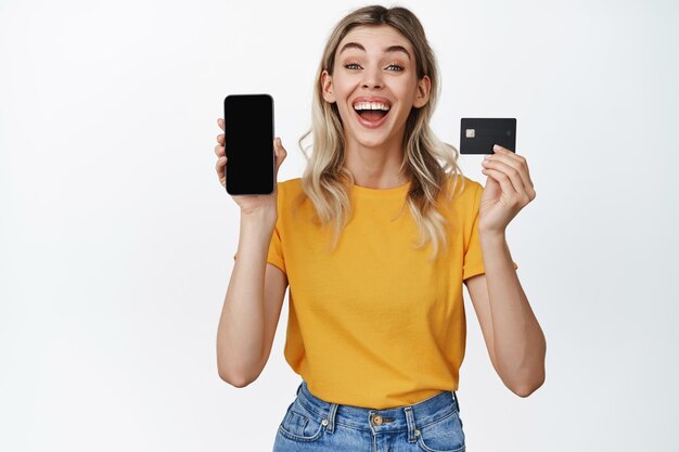 Retrato de una chica emocionada que muestra la pantalla del teléfono inteligente y la tarjeta de crédito que muestra la interfaz de la aplicación sobre fondo blanco