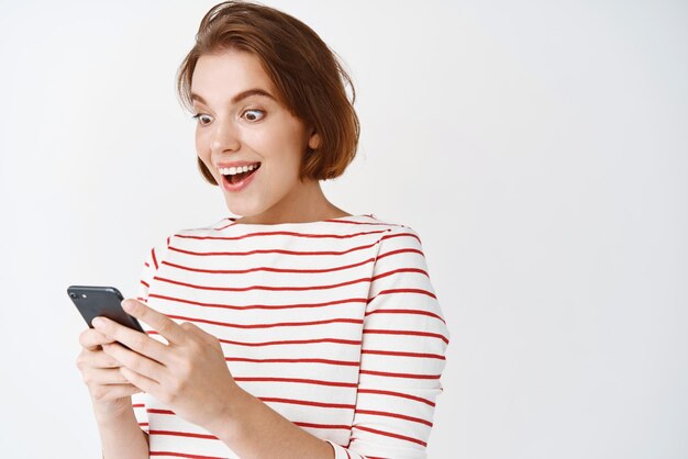 Retrato de una chica emocionada que se ve sorprendida por la pantalla del teléfono inteligente leyendo grandes noticias de pie feliz contra el fondo blanco