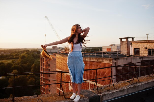 Retrato de una chica encantadora con ropa informal posando en el techo con el sombrero en la mano al atardecer