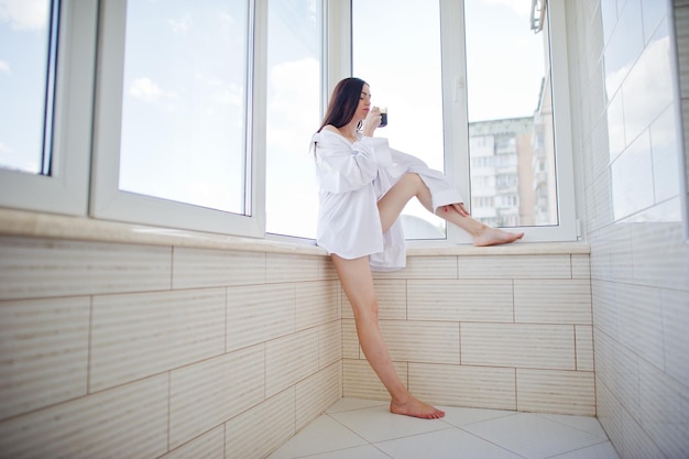 Retrato de una chica encantadora en ropa interior y camisa masculina de pie con un vaso de agua en las manos en el balcón