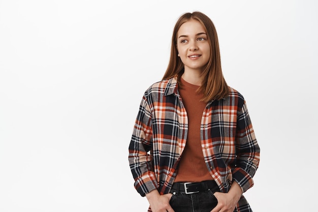 Foto gratuita retrato de una chica exitosa, una mujer elegante y segura de sí misma mirando a un lado con una expresión de cara sonriente y pensativa, tomándose de la mano en los bolsillos de los jeans, de pie contra un fondo blanco.