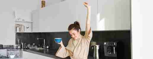 Foto gratuita retrato de una chica feliz bailando con café en la cocina con bata de baño disfrutando de su mañana