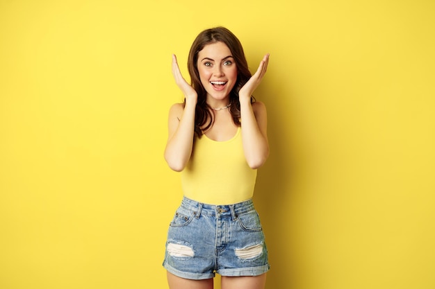 Foto gratuita retrato de una chica feliz con los ojos abiertos, mirando asombrada por la sorpresa y sonriendo, revelando un regalo, de pie con un traje de verano sobre un fondo amarillo.