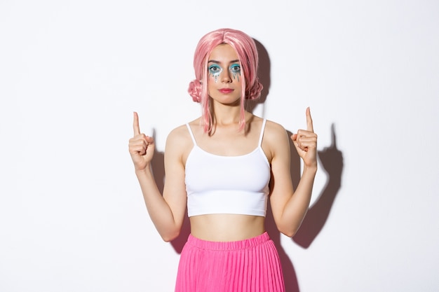 Retrato de chica fiestera con estilo con peluca de anime rosa y maquillaje de halloween, apuntando con el dedo hacia tu logo, de pie.