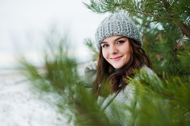 Retrato de una chica gentil con abrigo gris y sombrero contra el árbol de año nuevo al aire libre