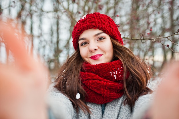 Retrato de una chica gentil con abrigo gris, sombrero rojo y bufanda cerca de las ramas de un árbol cubierto de nieve sosteniendo una cámara para selfie