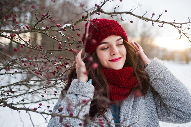 Retrato de una chica gentil con abrigo gris, sombrero rojo y bufanda cerca de las ramas de un árbol cubierto de nieve