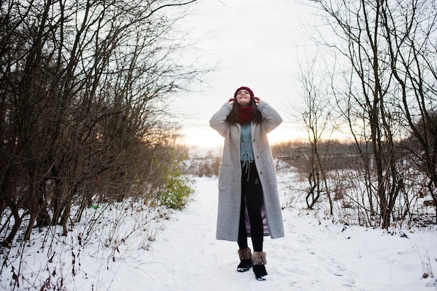 Retrato de una chica gentil con abrigo gris, sombrero rojo y bufanda cerca de las ramas de un árbol cubierto de nieve