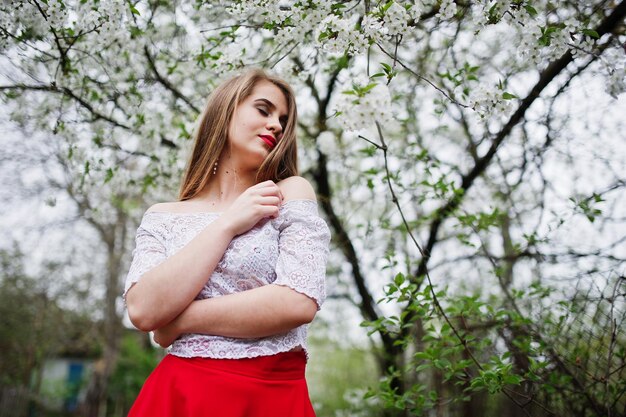 Retrato de una chica hermosa con labios rojos en el jardín de flores de primavera con vestido rojo y blusa blanca