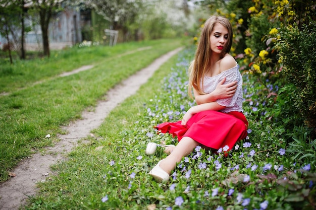 Retrato de una chica hermosa sentada con labios rojos en el jardín de flores de primavera en el césped con flores vestidas con vestido rojo y blusa blanca