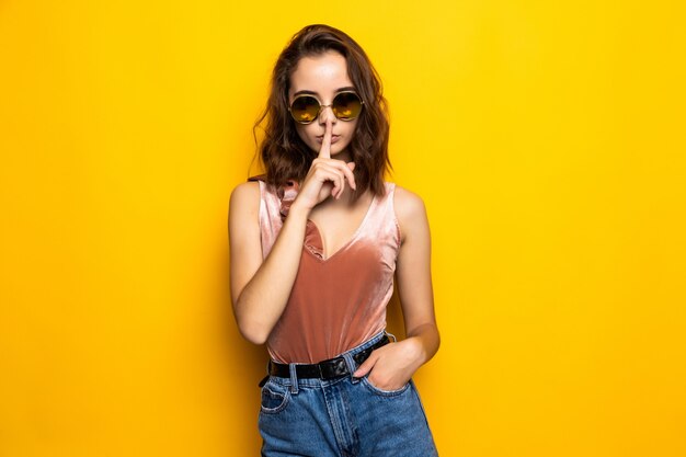 Retrato de una chica joven y bonita que muestra el gesto de silencio y un guiño aislado sobre amarillo