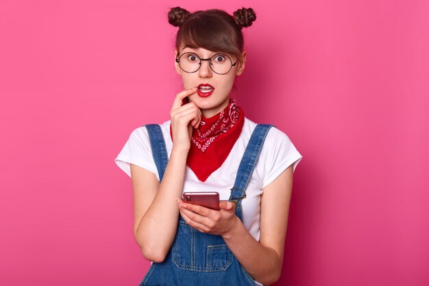 El retrato de una chica linda confundida usa un overol de mezclilla, una camiseta blanca informal, sostiene el teléfono móvil en la mano, mantiene el dedo índice en el labio