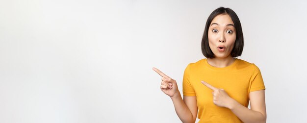 Retrato de una chica morena asiática sonriente con una camiseta amarilla que señala con el dedo a la izquierda mostrando un acuerdo promocional de espacio de copia que demuestra una pancarta de pie sobre fondo blanco