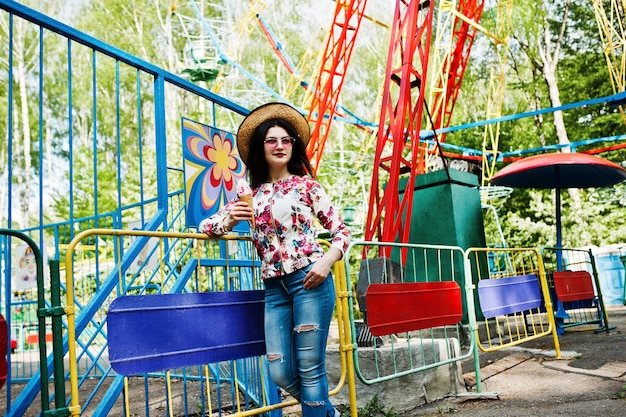 Retrato de una chica morena con gafas rosas y sombrero con helado en el parque de atracciones