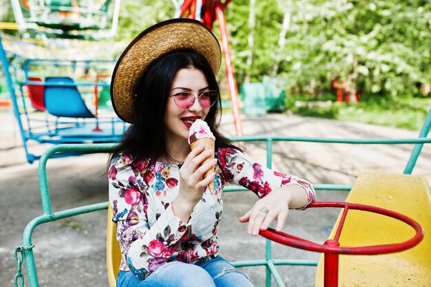 Retrato de una chica morena con gafas rosas y sombrero con helado en el parque de atracciones