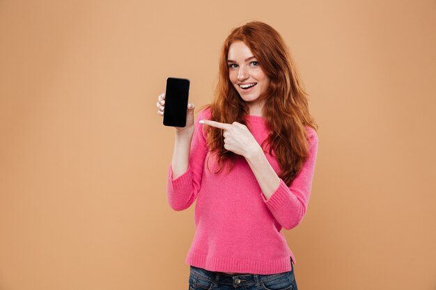 Retrato de una chica pelirroja atractiva emocionada apuntando al teléfono inteligente