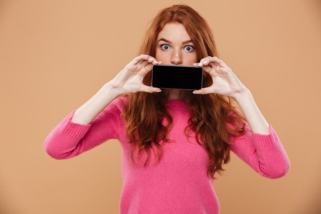 Retrato de una chica pelirroja atractiva sorprendida mostrando smartphone