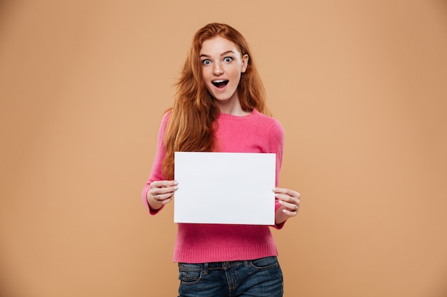 Retrato de una chica pelirroja bonita emocionada con cartel blanco en blanco