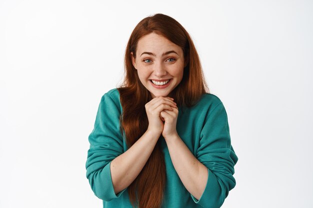 Retrato de una chica pelirroja esperanzada sonriendo y mirando con anticipación, esperando algo, rogando por un favor, de pie contra un fondo blanco.