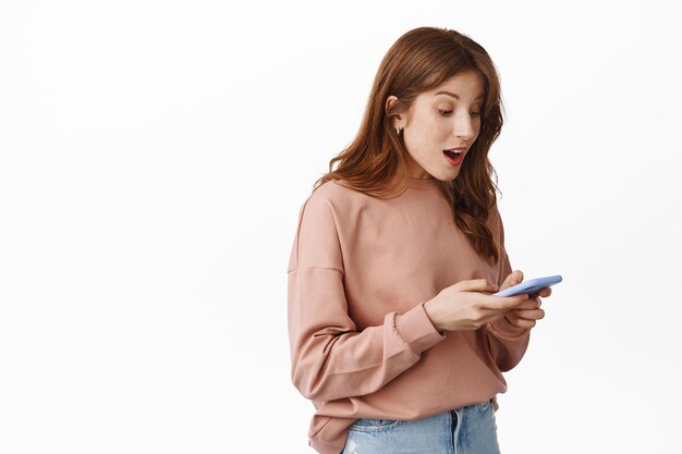 Retrato de una chica pelirroja sorprendida mirando la pantalla del teléfono con cara feliz, leyendo una notificación de mensaje en el móvil, chateando o comprando en línea, parada sobre fondo blanco