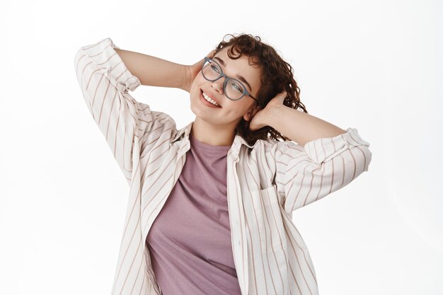 Retrato de una chica rizada relajada con gafas, sonriendo feliz, descansando y mirando la esquina superior izquierda, tomándose la mano en la cabeza sin preocupaciones, de pie contra el fondo blanco