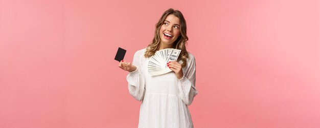 Retrato de una chica rubia atractiva y complacida con vestido blanco soñando con qué comprar con gi