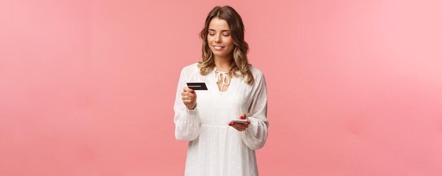 Retrato de una chica rubia atractiva y linda con un vestido blanco de peinado corto y rizado que sostiene números de dígitos de inserción de teléfono inteligente y tarjeta de crédito para registrarse en la aplicación de compras comprar fondo rosa en línea