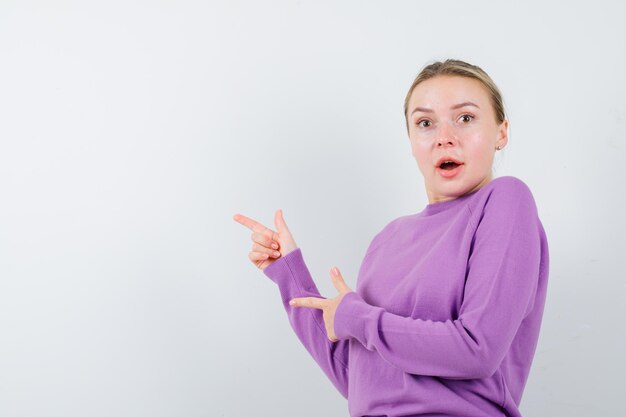 Retrato de una chica rubia atractiva muy sorprendida en un suéter morado sorprendentemente apuntando hacia la izquierda aislado sobre fondo blanco.