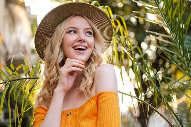 Retrato de una chica rubia bastante alegre con sombrero felizmente mirando hacia otro lado en la calle de la ciudad