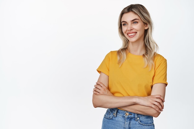 Retrato de una chica rubia confiada sonriendo con los brazos cruzados en el pecho y mirando a un lado de pie con camiseta amarilla y fondo blanco de jeans