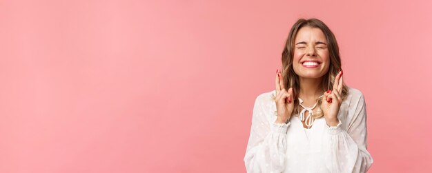 Retrato de una chica rubia emocionada y esperanzada que desea cruzar los dedos para tener buena suerte, cerrar los ojos y sonreír poniendo todo su esfuerzo en rezar suplicando que el sueño se haga realidad anticipando el fondo rosa