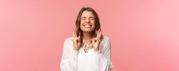 Retrato de una chica rubia emocionada y esperanzada que desea cruzar los dedos para tener buena suerte, cerrar los ojos y sonreír poniendo todo su esfuerzo en rezar suplicando que el sueño se haga realidad anticipando el fondo rosa