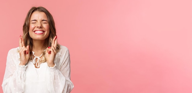 Retrato de una chica rubia emocionada y esperanzada que desea cruzar los dedos para tener buena suerte, cerrar los ojos y sonreír poniendo todo su esfuerzo en rezar suplicando que el sueño se haga realidad anticipando el fondo rosa